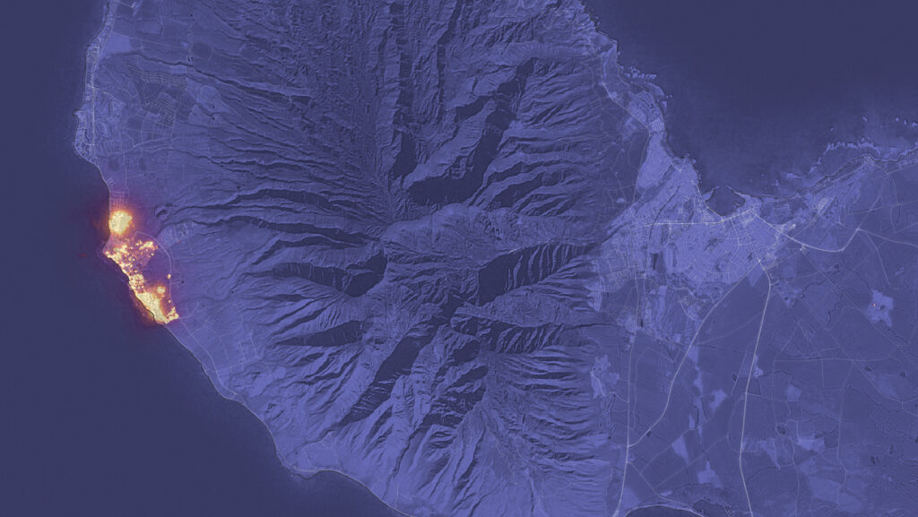 A night scene shows a bright orange patch near the coast burning the city of Lahaina. The rest of the land looks purple in this night image.
